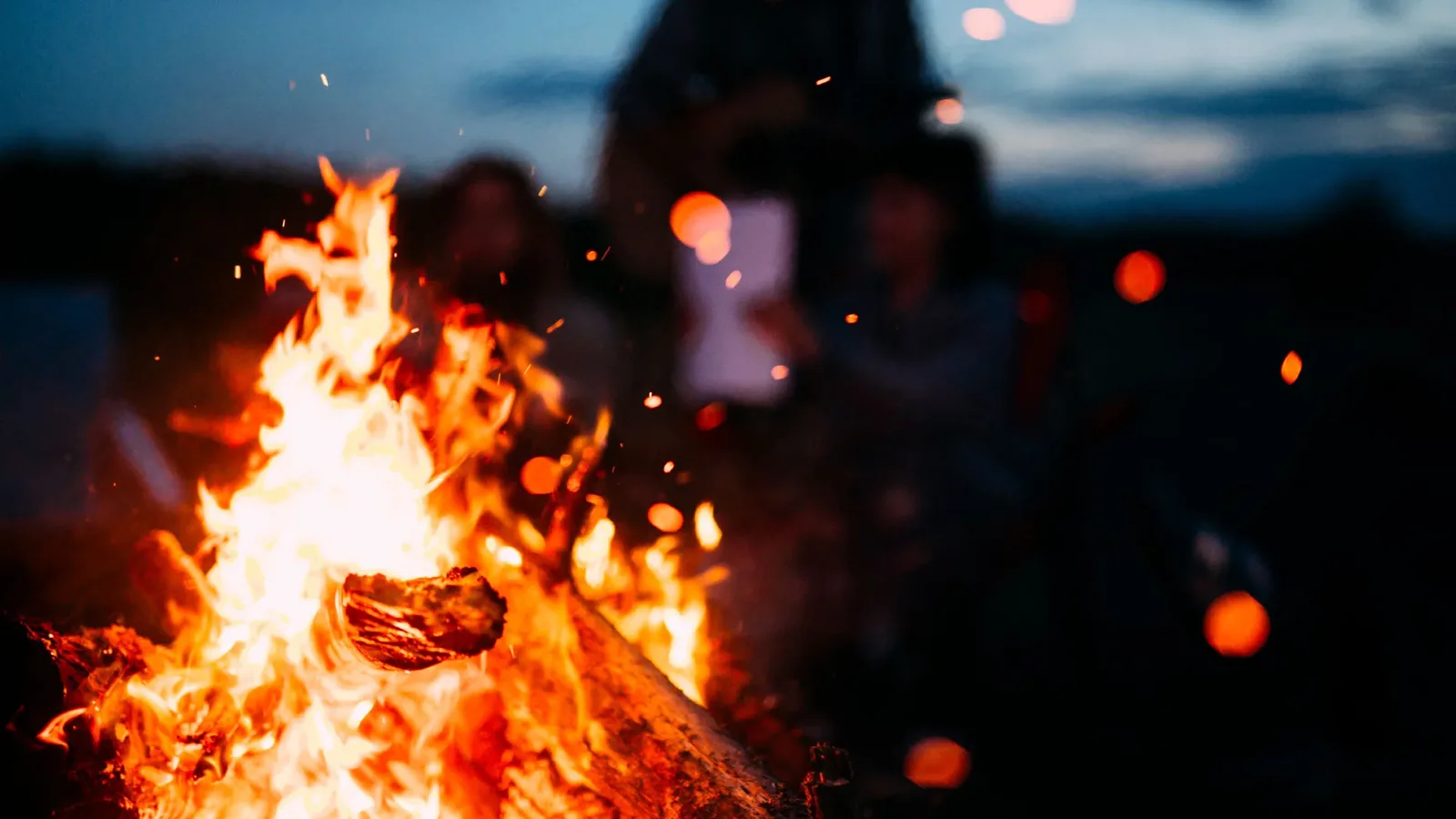 A wood fire burning at night, sparks rising, people gathered out of focus behind it