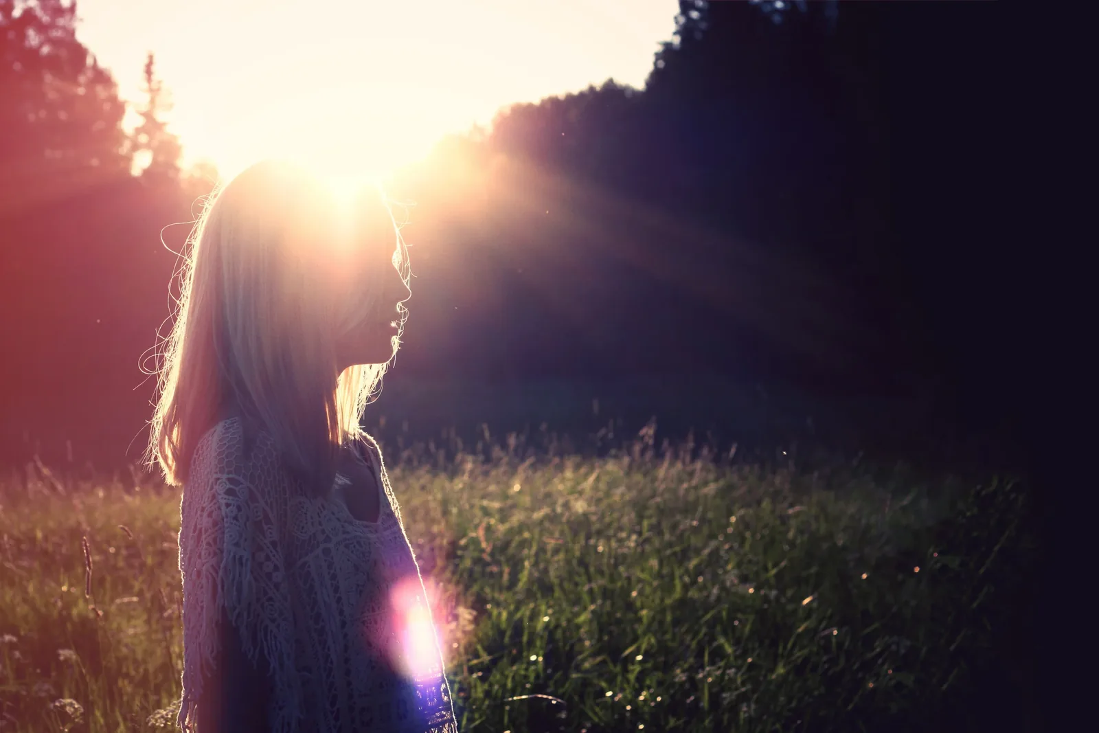 A woman standing in tall grass, facing the low sun through the trees