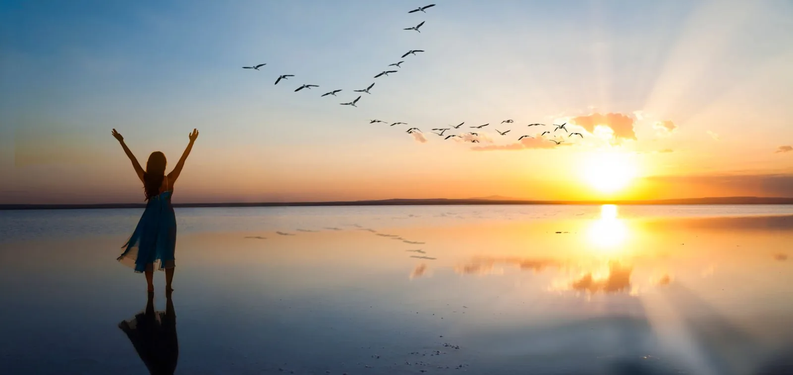 A woman with her arms raised on a beach at sunrise, birds flying overhead