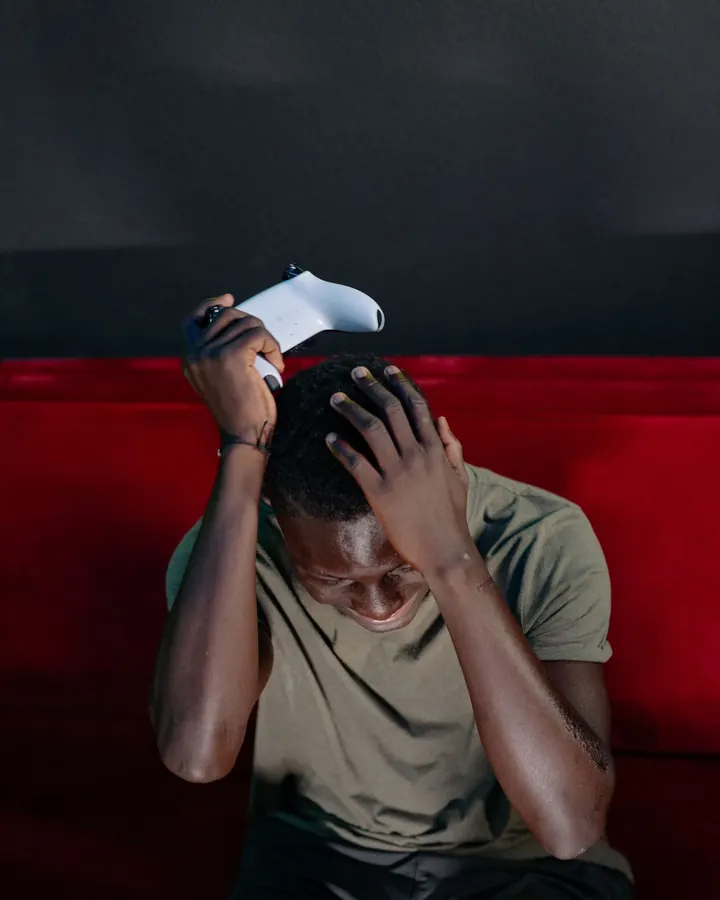 A young man on a couch with both hands on his head, a game controller in one hand, face hidden