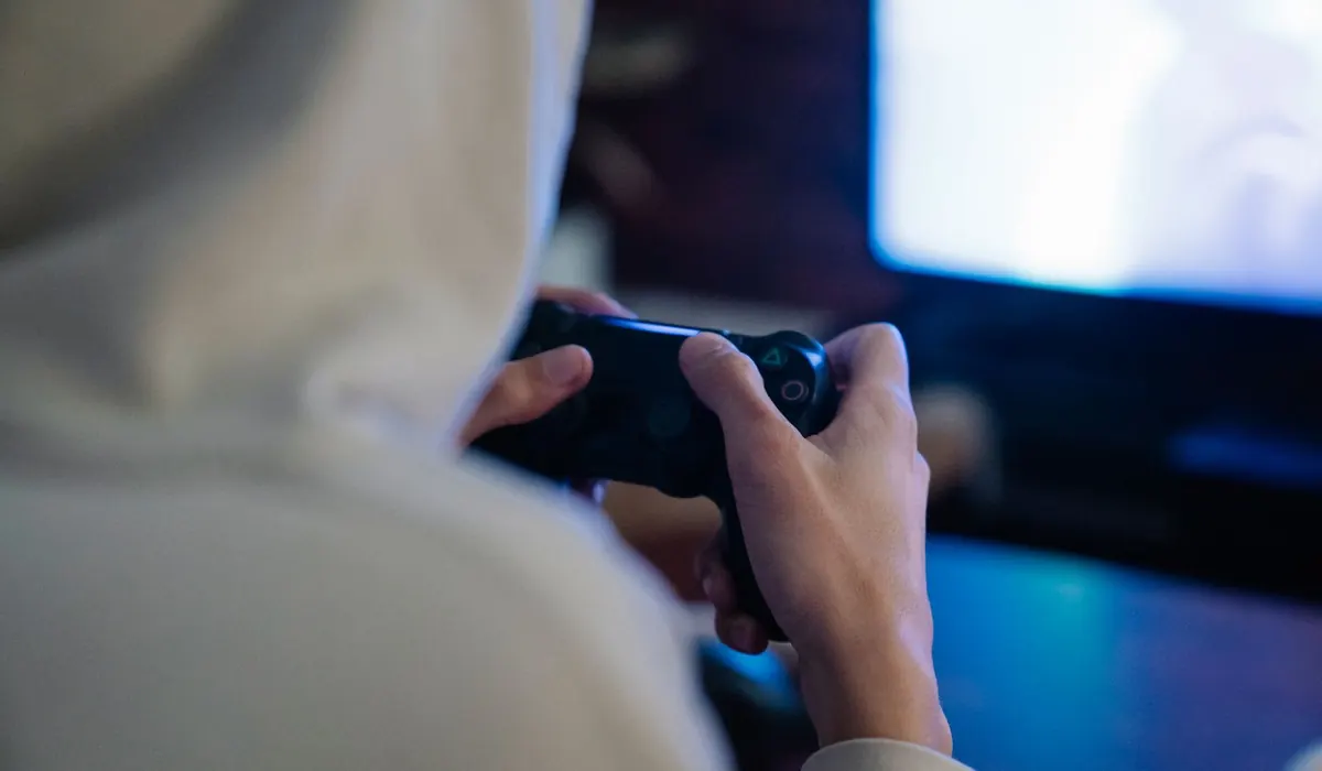 A player in a hooded top, seen from behind, holding a controller in front of a glowing screen in a dark room